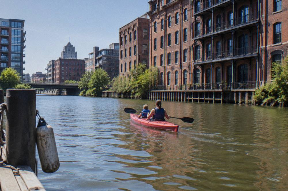 Tandem kayaks floating on a calm downtown river stretch with brick buildings behind