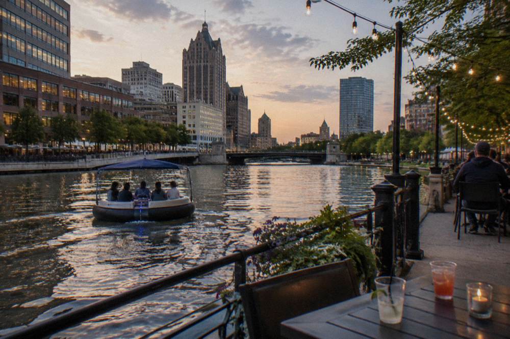 A round electric donut boat cruising a downtown river beside a riverside walkway