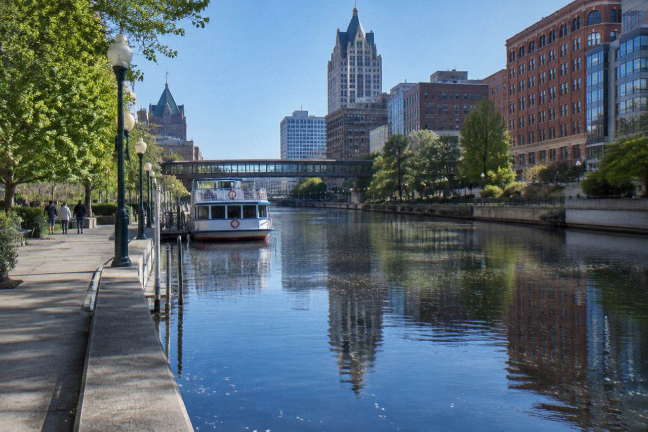 A calm stretch of the Milwaukee River with downtown buildings and the Riverwalk on a clear day