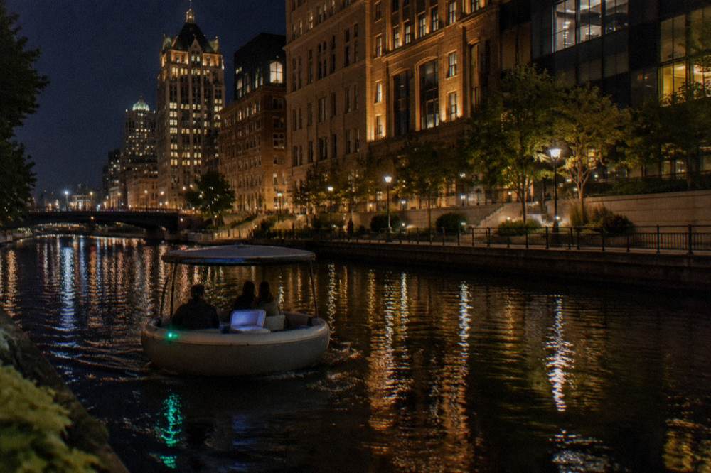 A small round electric boat cruising past lit-up buildings on a downtown riverwalk at night