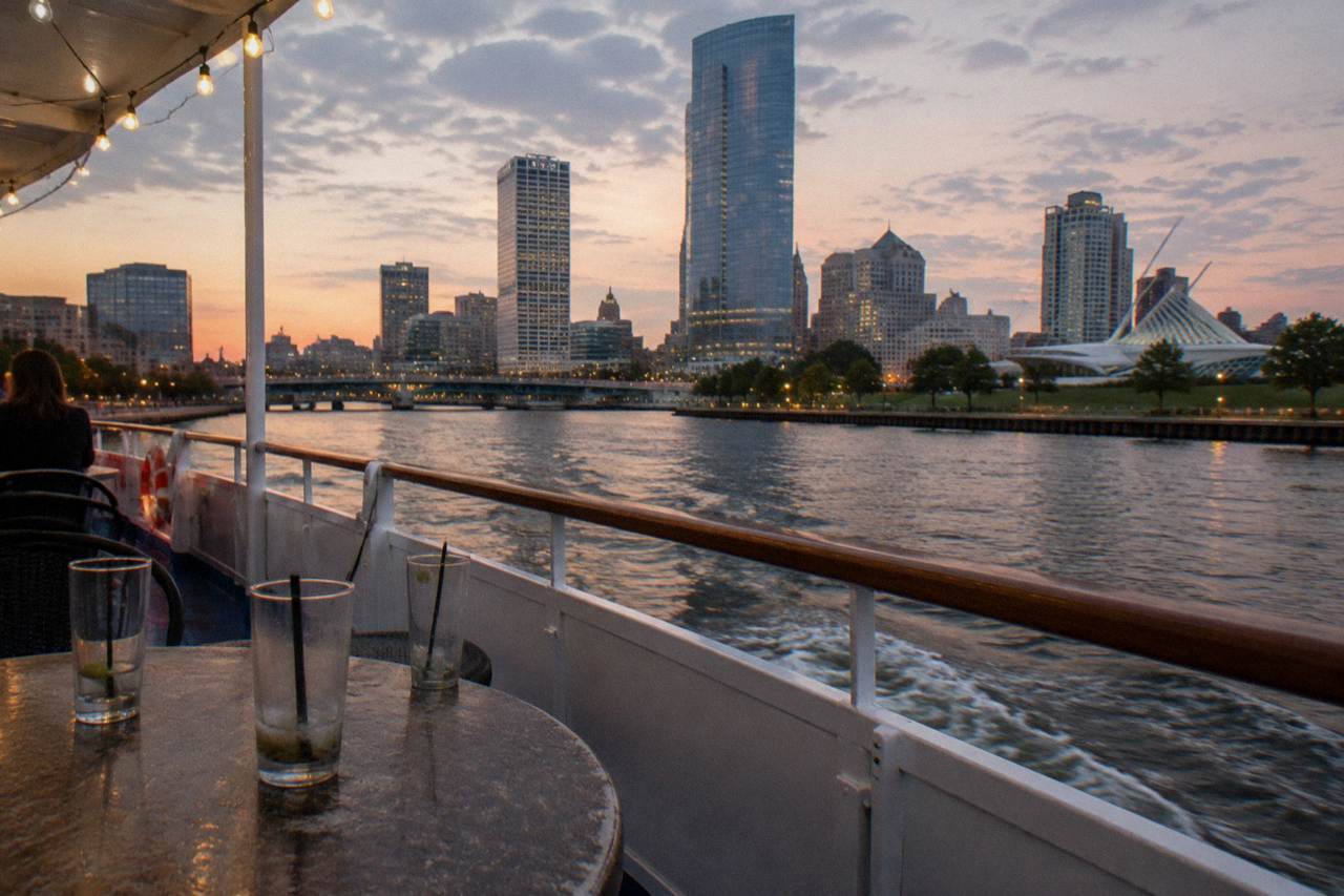 A cruise boat deck on the Milwaukee River at dusk with the downtown skyline behind it