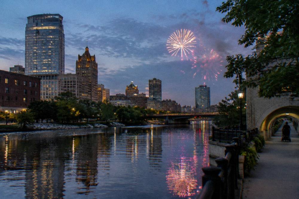 A city skyline at dusk reflected in a calm river with a riverside walkway