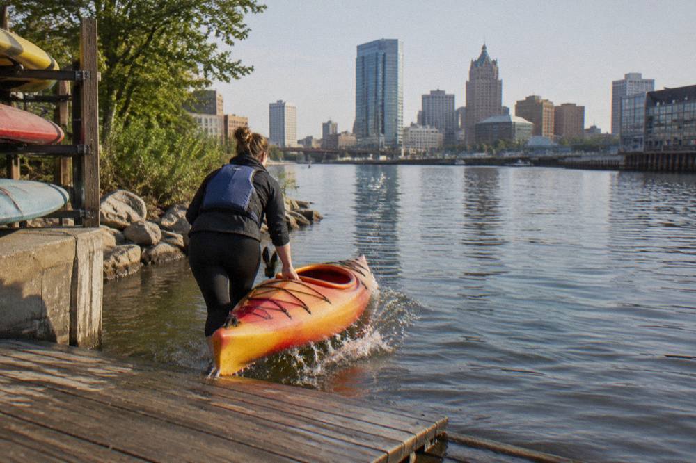 A kayak slipped into the Milwaukee River from a wooden launch dock with the downtown skyline in the background