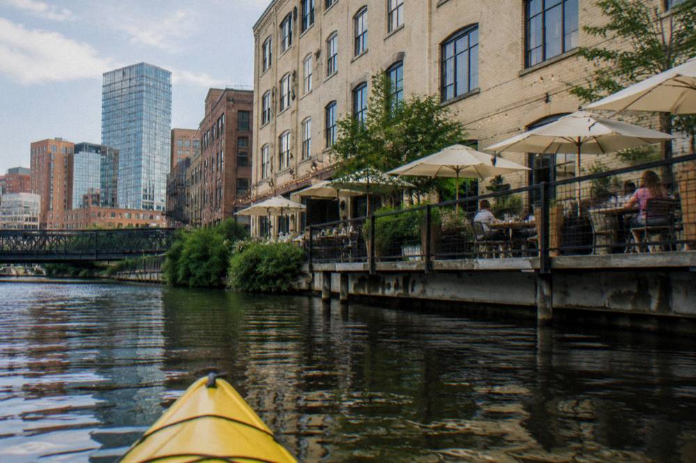 A kayak floating near the Riverwalk with restaurant patios and downtown buildings reflected in the river