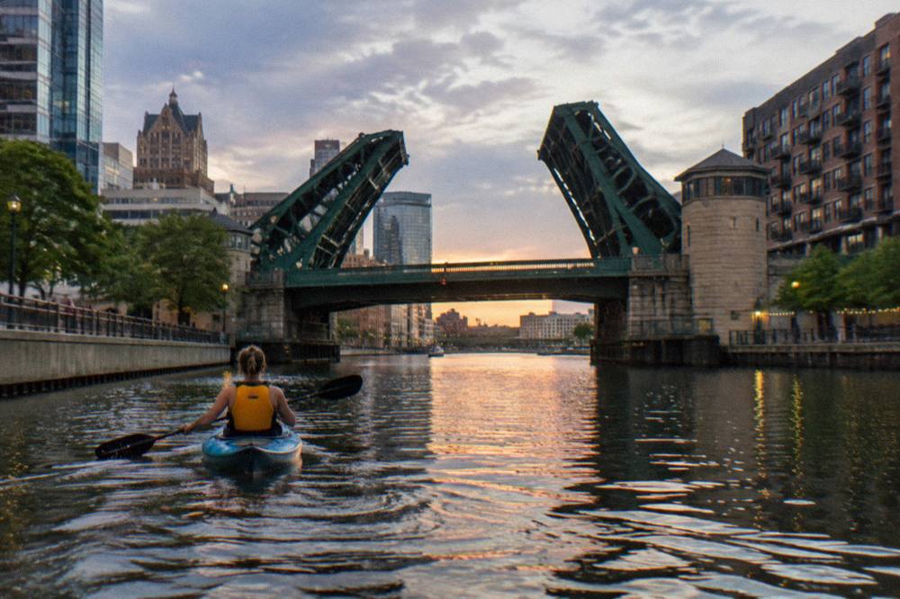 A paddler in a life jacket gliding under a downtown Milwaukee bascule bridge on calm evening water