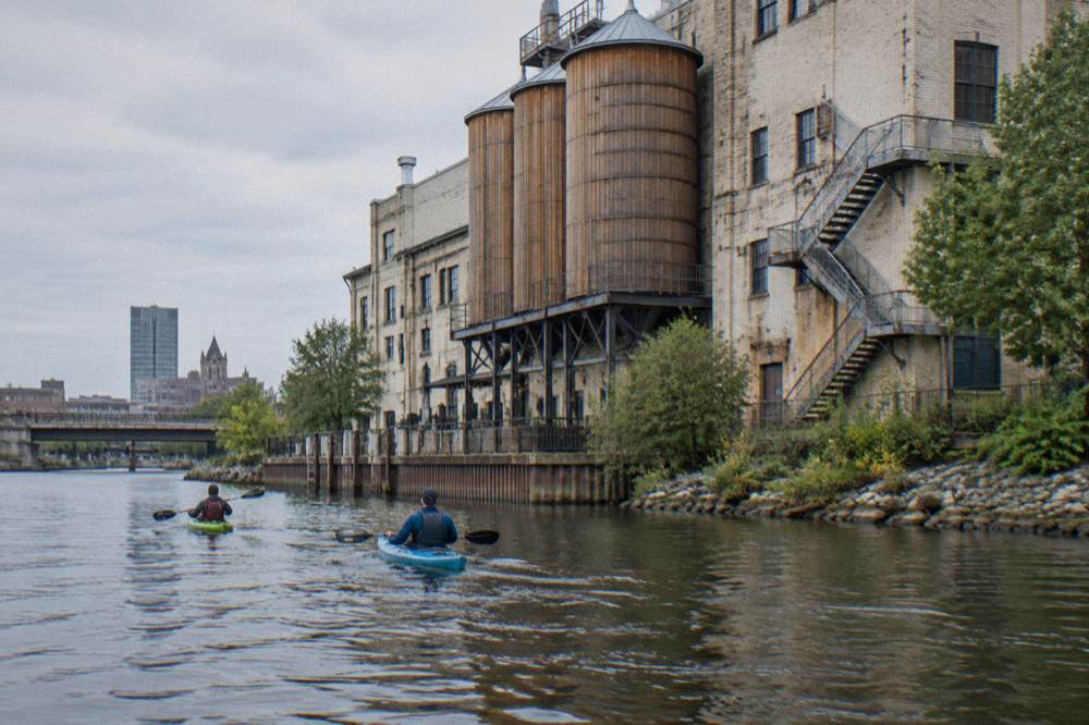 Two kayakers paddling past brewery buildings along the Milwaukee River