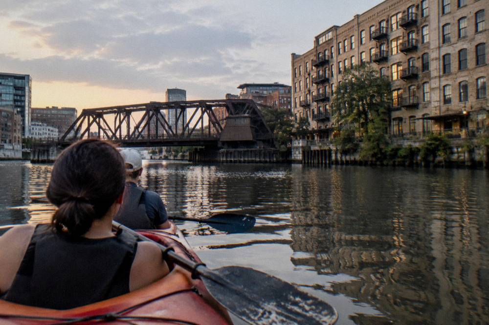 A tandem kayak floating on calm river water beside a downtown Milwaukee bridge