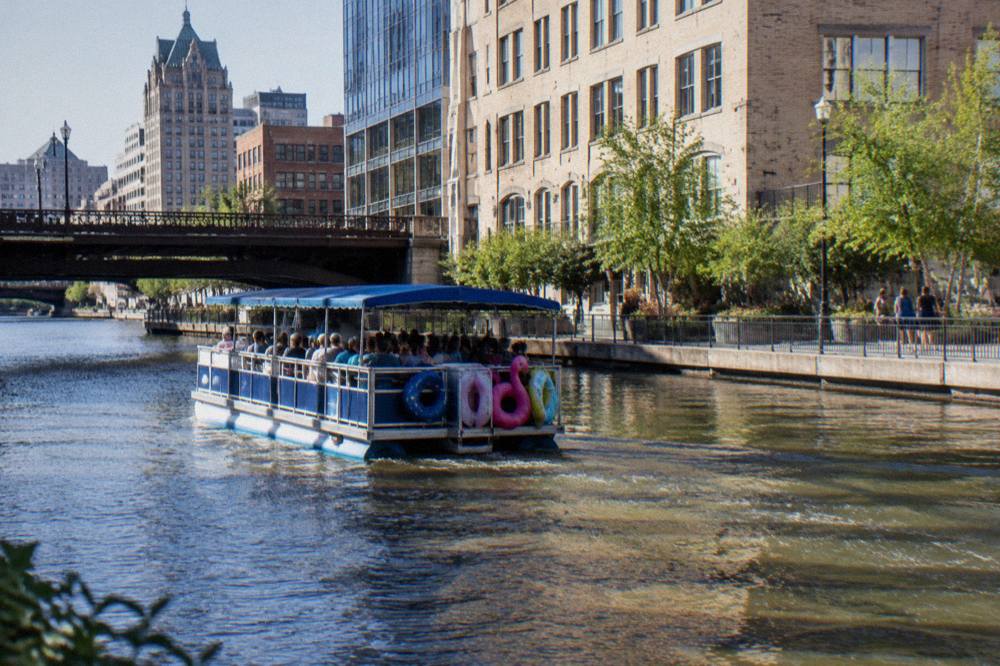 A tour boat moving down the Milwaukee River between downtown buildings on a bright afternoon