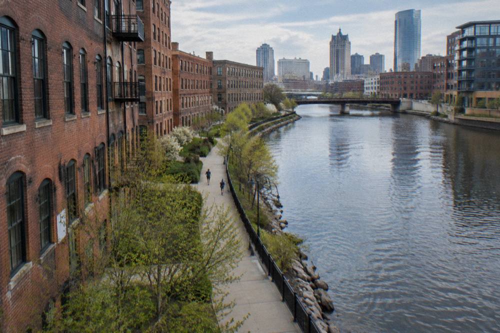 Riverside walkway in front of brick warehouse buildings with the city skyline behind