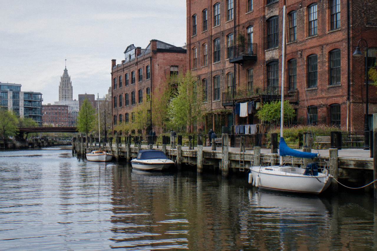 Red brick warehouse buildings along the Milwaukee River with a riverside walkway and boats at dock