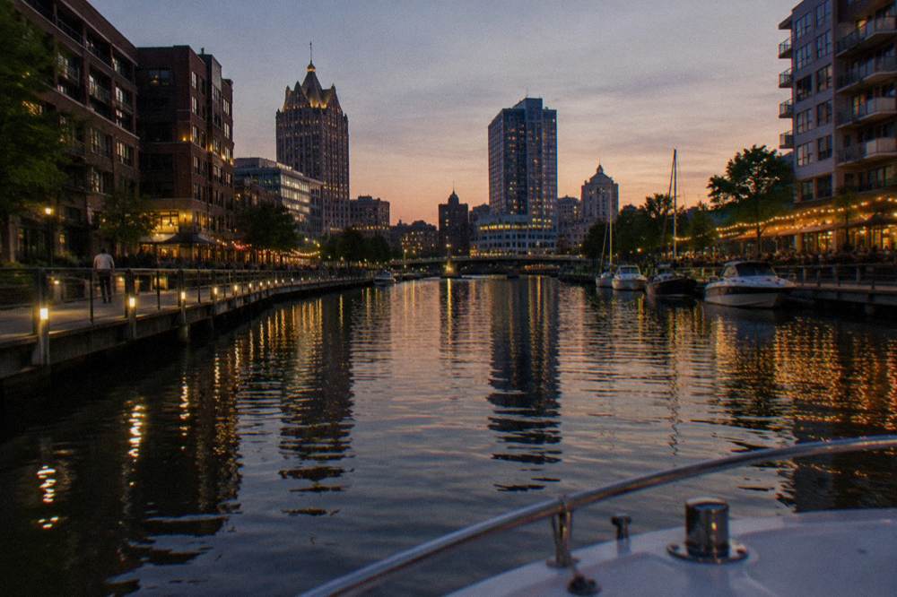 Boats tied along the Milwaukee Riverwalk at dusk with lit restaurant patios on both banks
