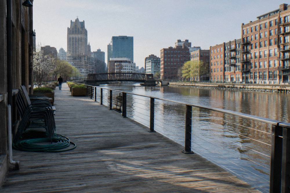 Wide Third Ward boardwalk along the Milwaukee River with timber decking and buildings on the far bank