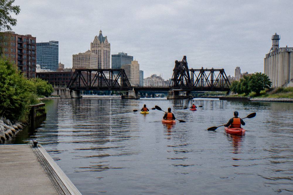 Paddlers in life jackets crossing the calm Milwaukee River between downtown buildings